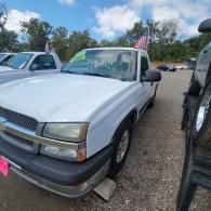 Silver pickup truck parked outdoors between two vehicles, with an American flag visible in the background.