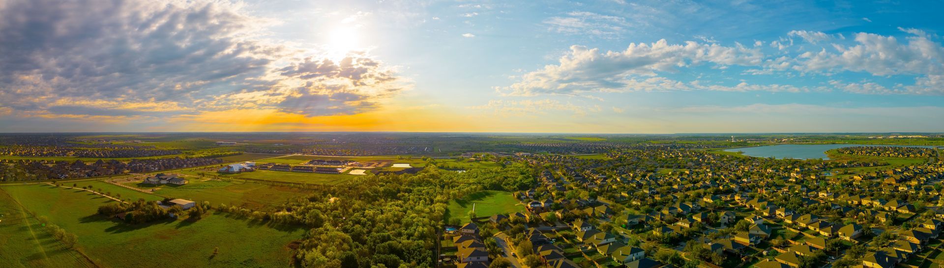 Aerial view of a town with green trees and a lake under a cloudy sky at sunset.