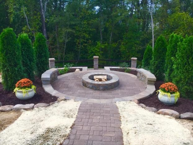 A circular stone patio with a central fire pit, curved seating walls, and potted orange mums, framed by tall evergreen trees.