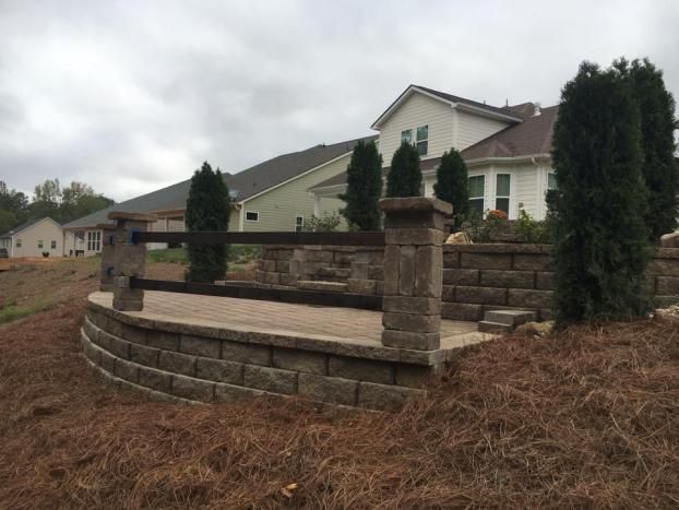 A tan stone patio with a low block retaining wall, capped stone pillars, and dark wood railings, set in a grassy yard.