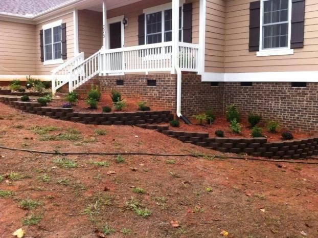 A tan house with dark shutters and a brick foundation, featuring a newly installed tiered stone retaining wall garden.
