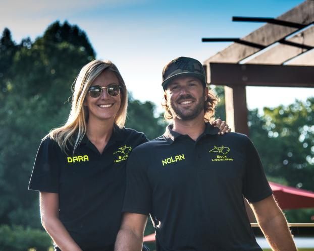A woman and a man wearing matching black shirts with names and logos, standing side-by-side outdoors under a trellis.