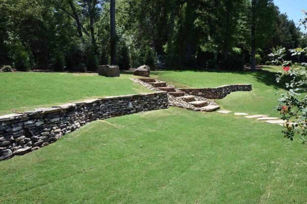 A stone retaining wall curves across a green lawn, with stone steps leading up a slight incline toward trees in the distance.