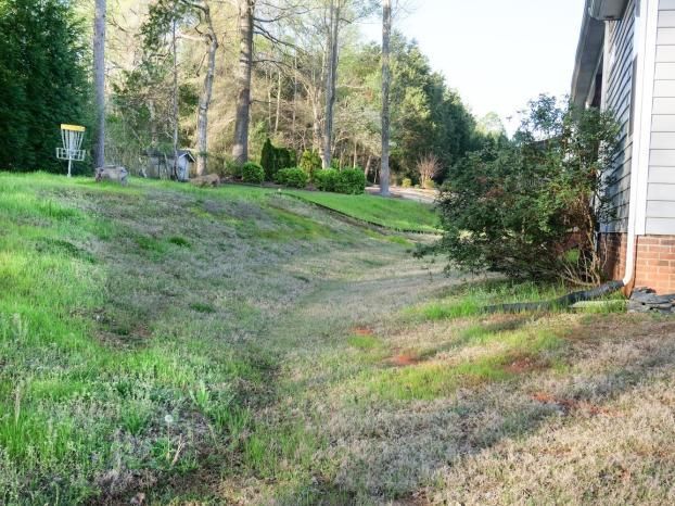 A grassy, shallow drainage swale leads from the side of a residential house toward a wooded area with a disc golf basket.