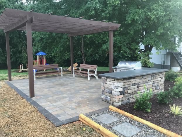 A stone outdoor kitchen with a grill next to a wooden pergola and two benches on a paved patio in a grassy backyard.