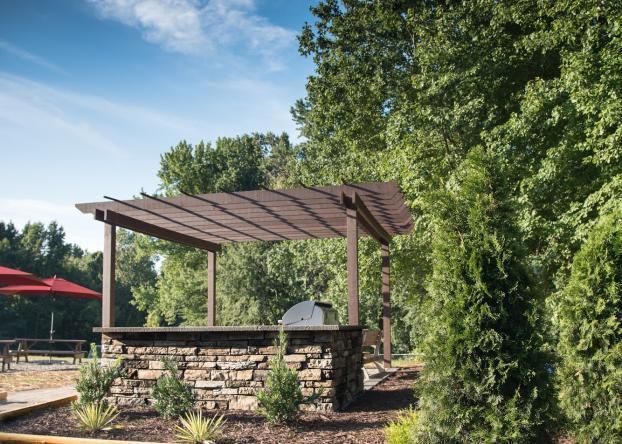 A stone outdoor kitchen with a wooden pergola set in a lush, wooded backyard.