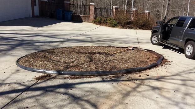 A circular garden bed filled with brown mulch sits in the middle of a concrete driveway, next to a gray pickup truck.