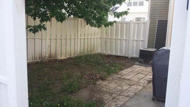 A small backyard with a patch of dirt and grass, a light-colored stone patio, a white vinyl fence, and a covered grill.
