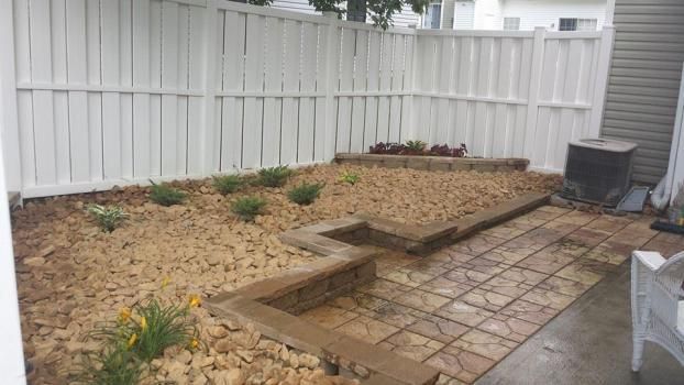 A small backyard with a paved stone patio, a tiered retaining wall, and gravel garden beds enclosed by a white fence.