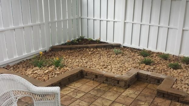A patio featuring tan pavers, a low brick retaining wall, a gravel garden bed with small plants, and a white privacy fence.