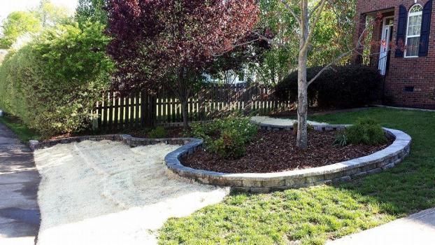 A landscaped front yard features a curved stone retaining wall surrounding mulch, trees, and shrubs beside a gravel path.