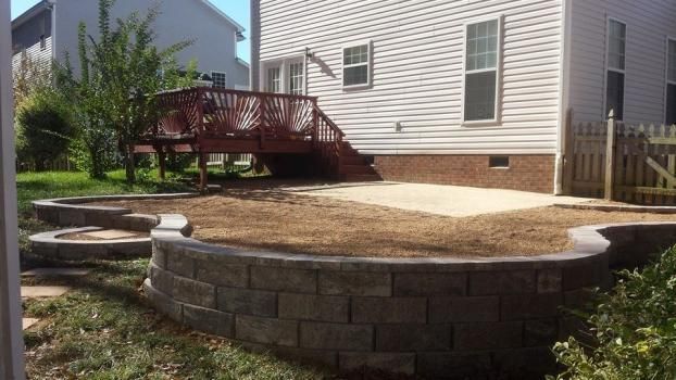 A backyard landscape featuring a curved stone retaining wall, a patch of dirt, a concrete patio, and a red wooden deck.