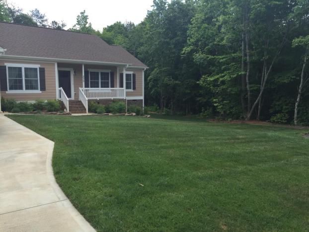 A tan, single-story house with a brown roof and a front porch, viewed from a paved driveway next to a grassy lawn.