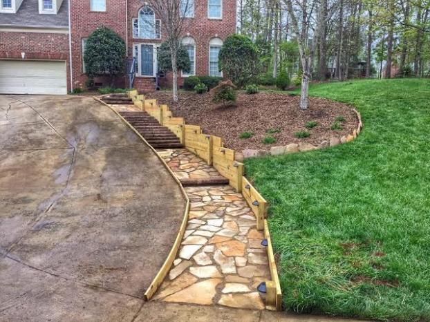 A stone walkway framed by a wood retaining wall next to a concrete driveway and a grassy slope leading to a brick house.