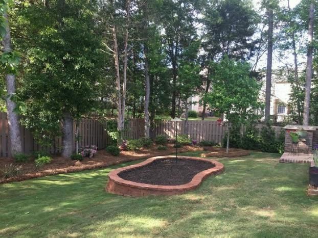 A backyard garden area featuring a raised, kidney-shaped brick planter filled with dark soil, surrounded by lawn and trees.