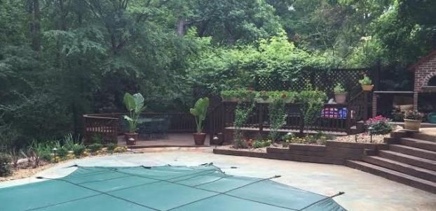 A backyard patio with a covered swimming pool, tiered stone stairs, potted plants, and lush green trees in the background.