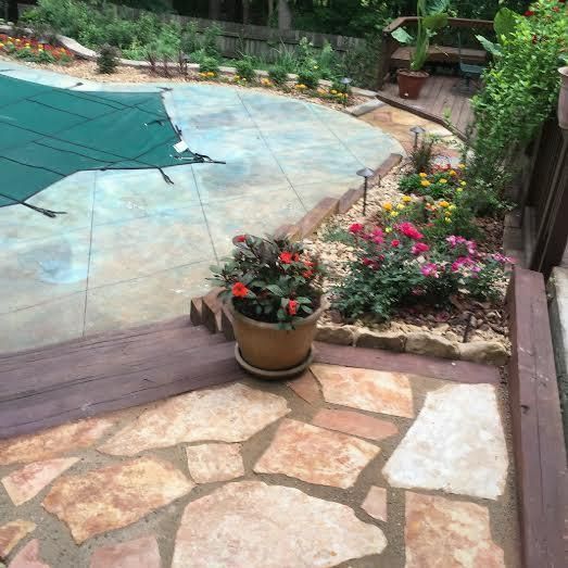 A stone patio and wooden deck lead to a backyard swimming pool covered by a green tarp, surrounded by flower beds.
