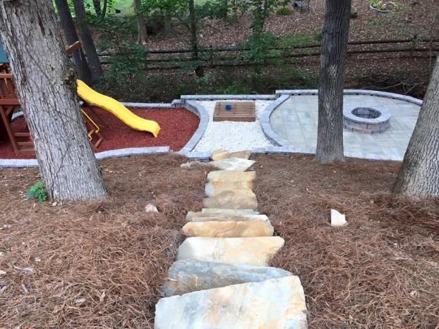 Stone stairs lead down a mulched slope to a backyard featuring a red play area with a yellow slide and a stone fire pit.