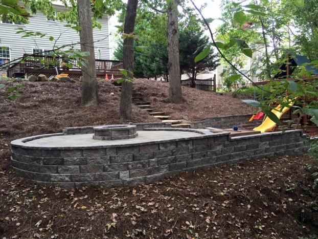 A tiered backyard featuring a stone patio with a built-in fire pit, stone retaining walls, and a yellow slide.