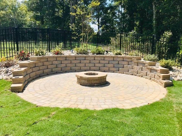 A circular stone patio with a central fire pit, enclosed by a curved retaining wall in a grassy backyard.