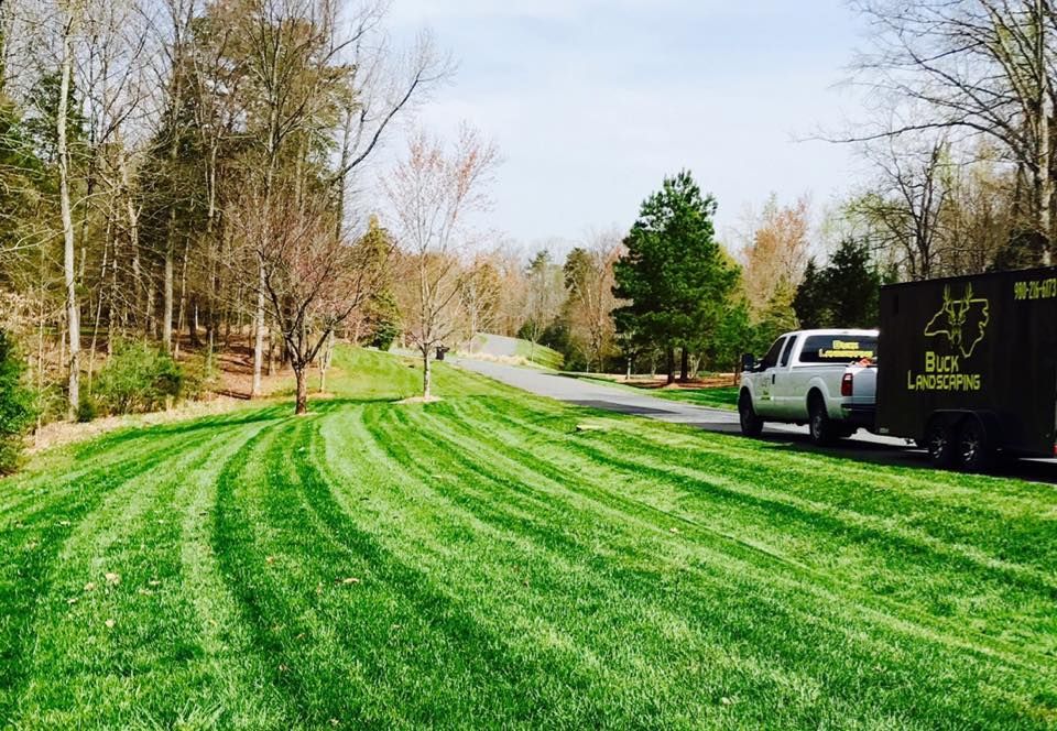 A white landscaping truck pulling a trailer is parked on the side of a road next to a lawn with fresh, curved mow lines.