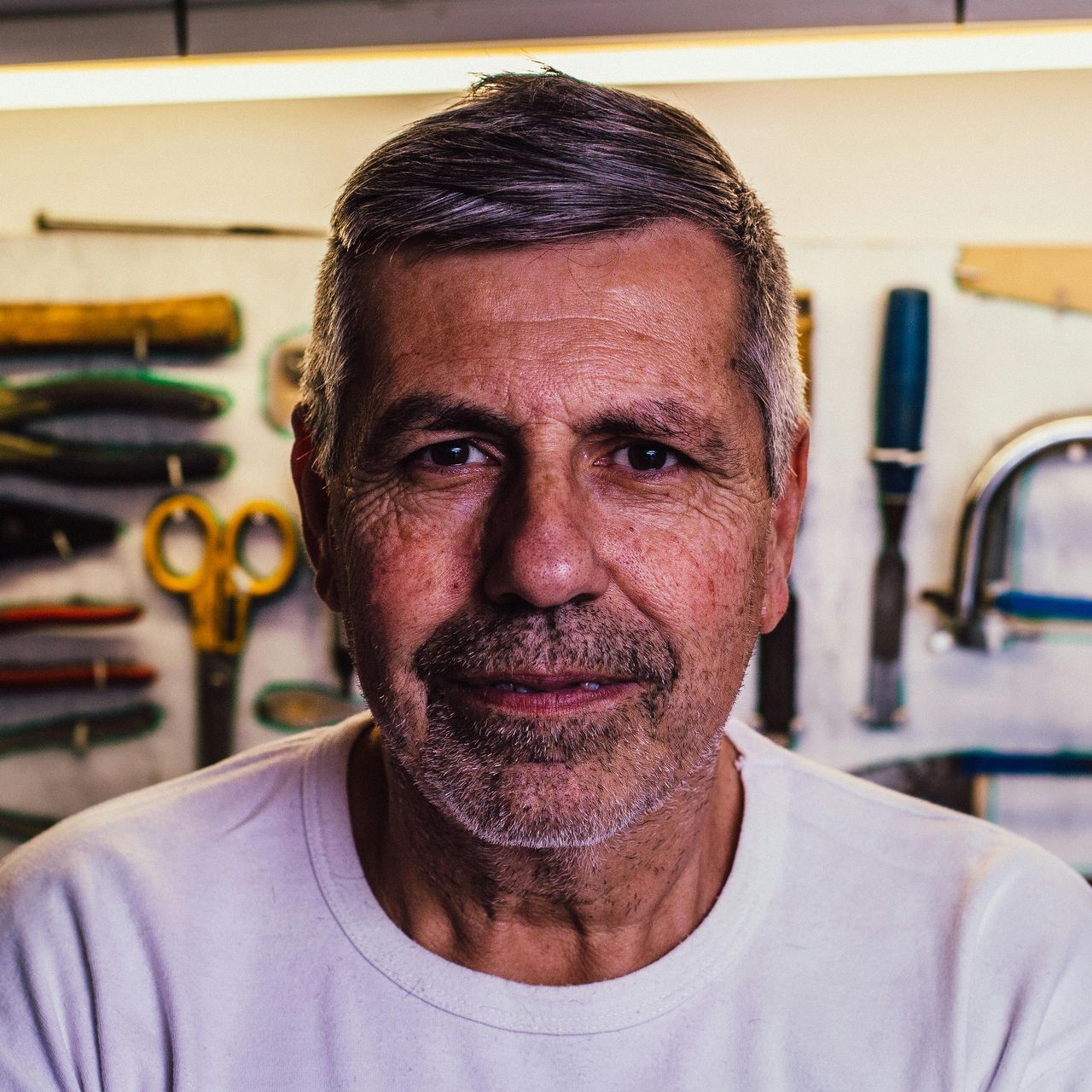 A man stands in a workshop with assorted tools visible on a wall board behind him.