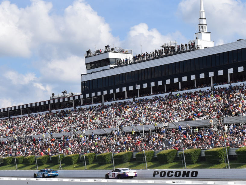 Two cars are racing on the Poconos race track in front of a crowd.