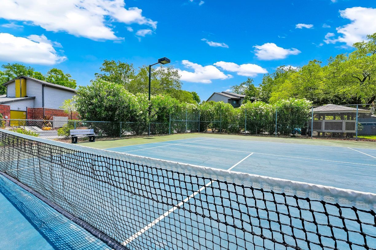 Tennis court with net in the foreground, blue surface, greenery, and buildings under a bright blue sky.