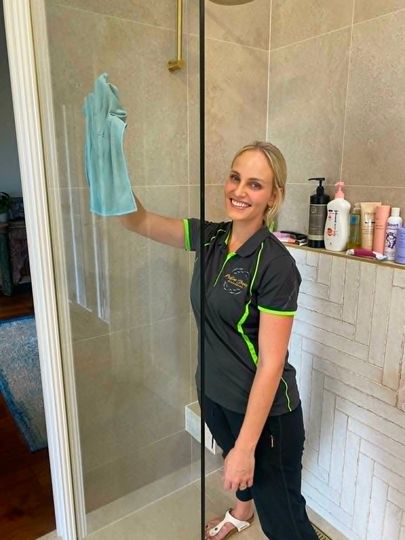 Woman cleaning a glass shower door in a bathroom, holding a blue cloth and smiling.