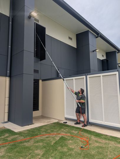 Person washing an upper-story window with a long pole beside a modern house and lawn