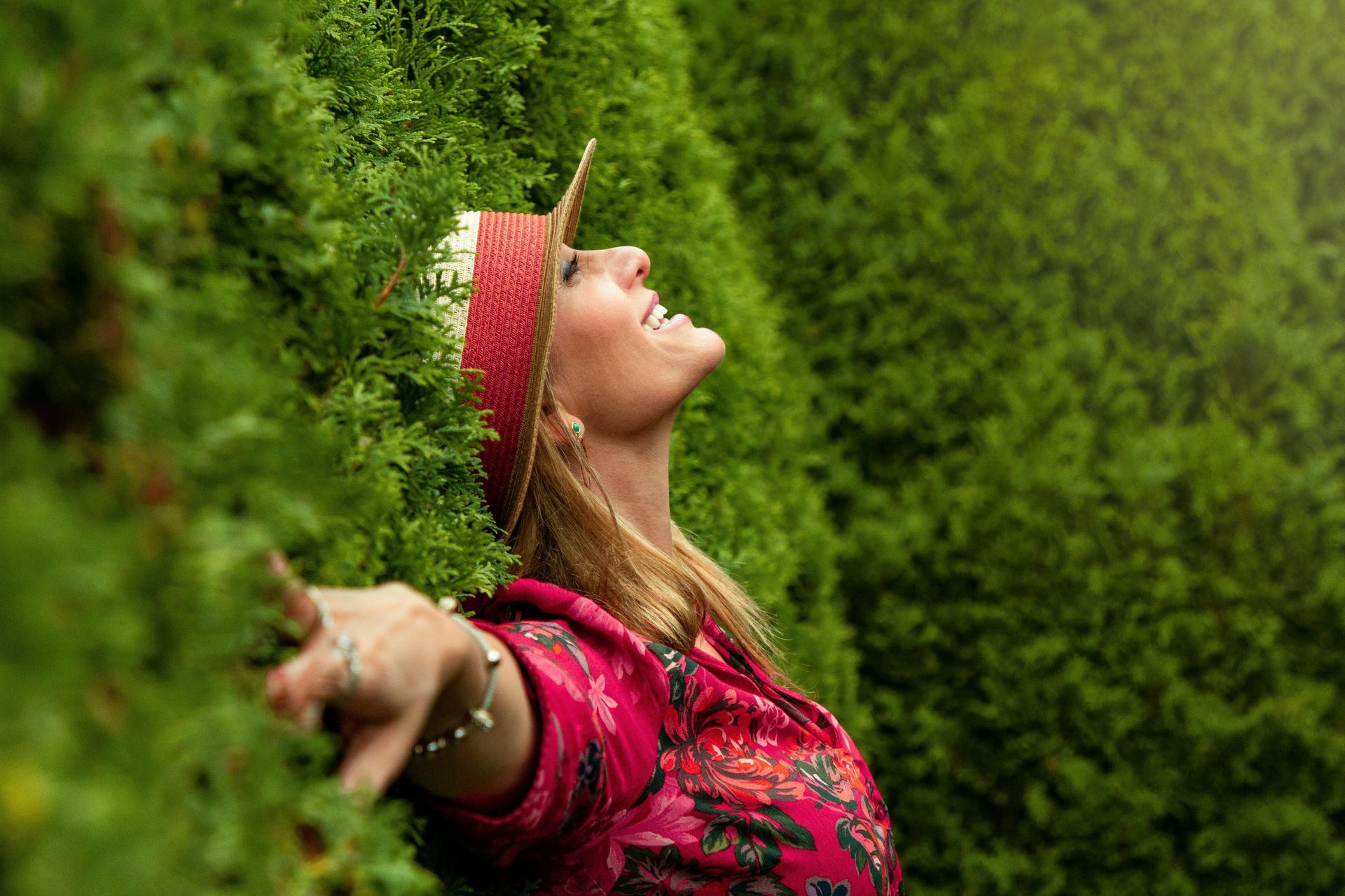 Woman in a red shirt and hat with arms outstretched, smiling, facing upwards near a green hedge.