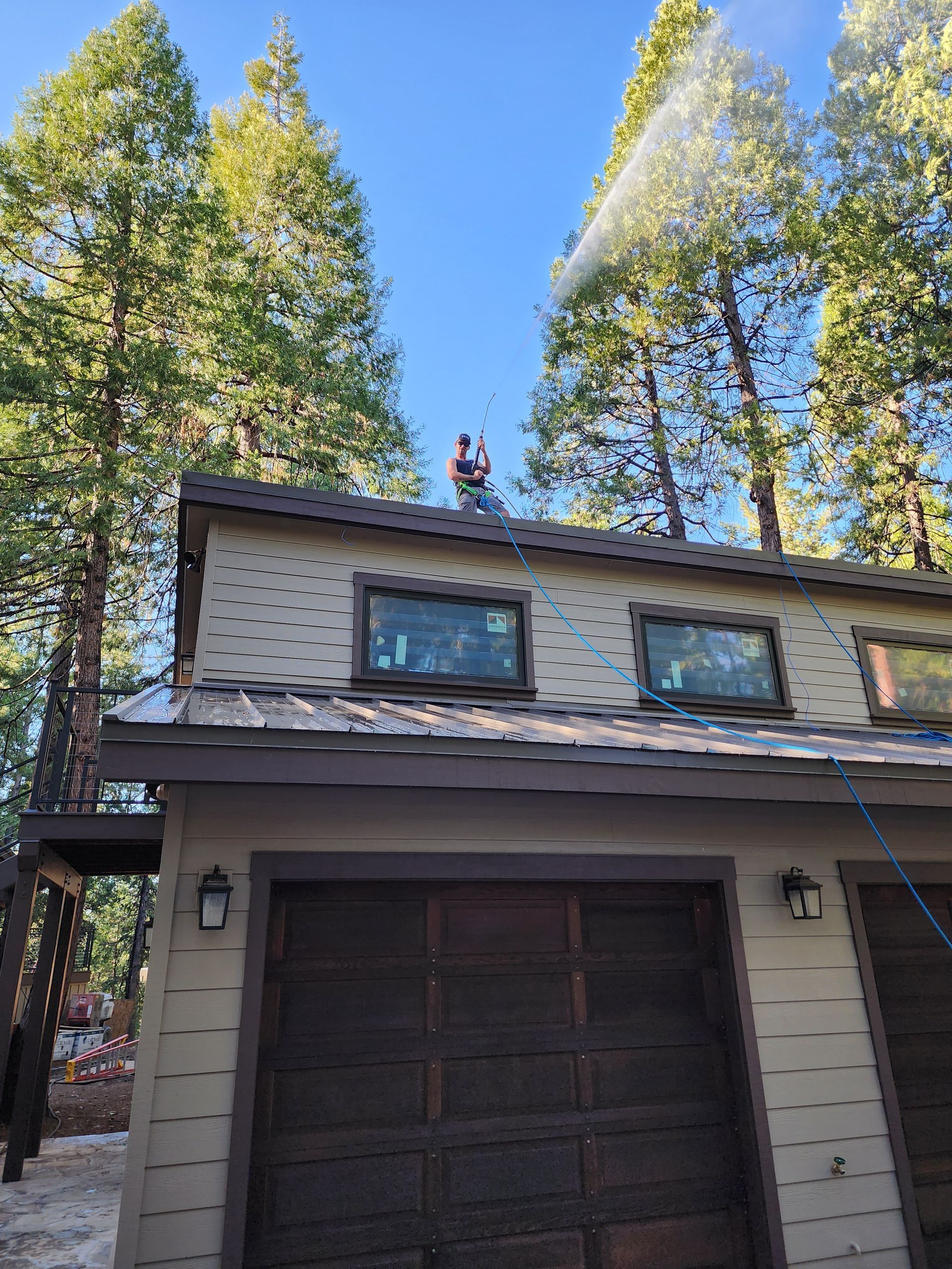 A man is spraying water on the roof of a house surrounded by trees.