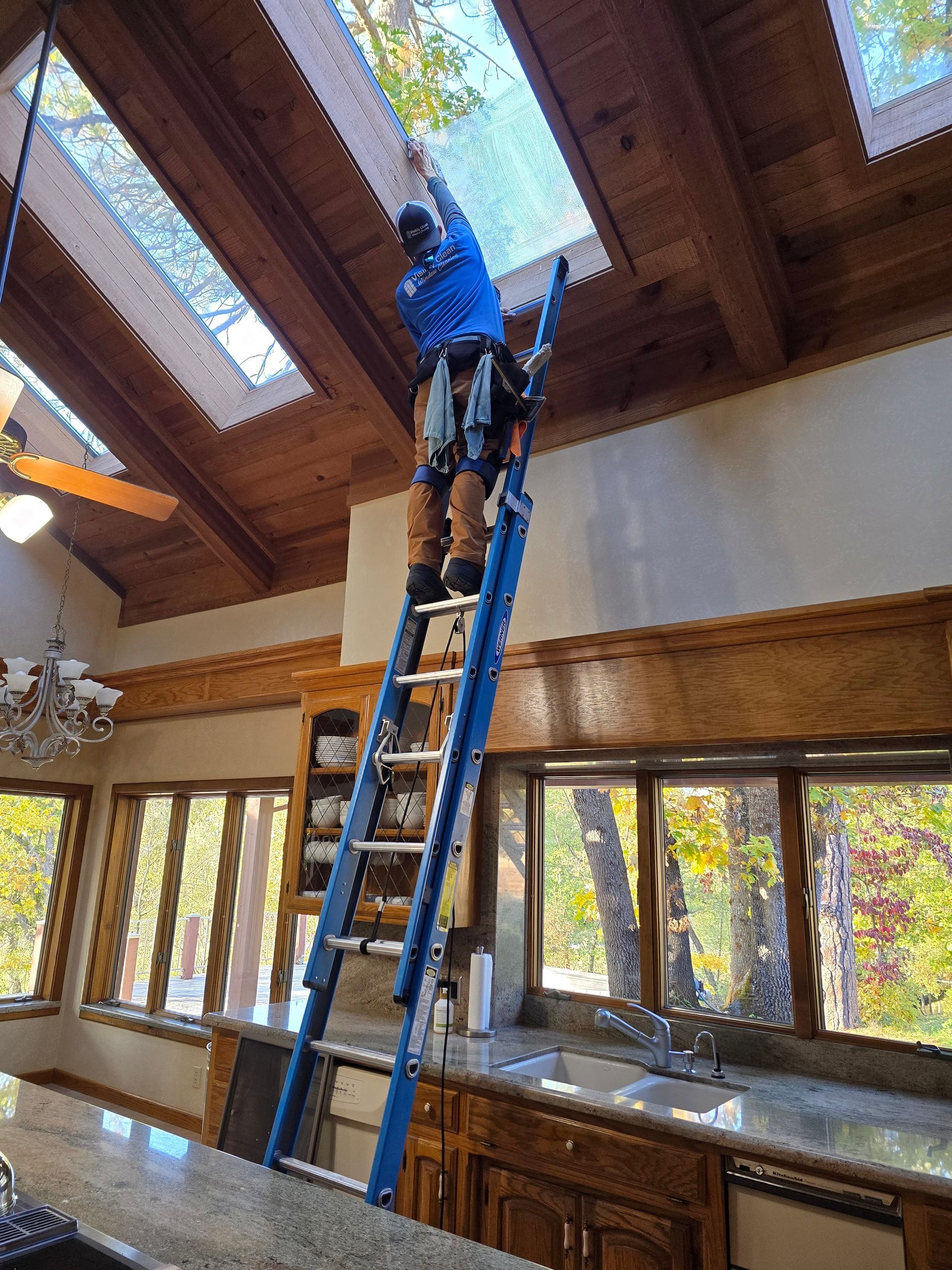 A man is standing on a ladder in a kitchen cleaning a skylight.