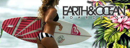 A woman in a bikini is carrying a surfboard in front of a sign that says earth & ocean board shop