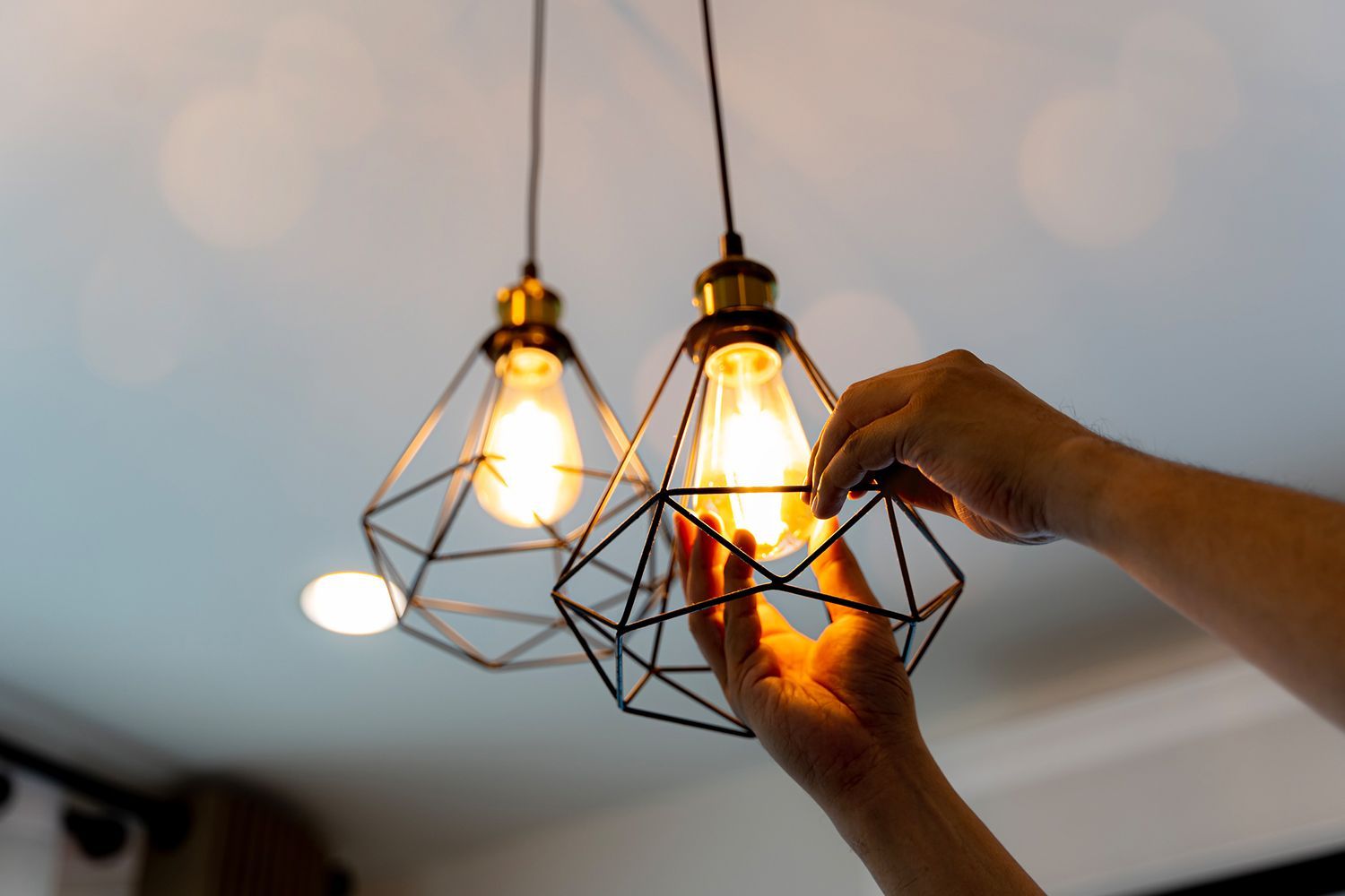 Person's hands adjusting a hanging pendant light with a geometric cage design, near another similar light on a white ceiling. Person's hands adjusting a hanging pendant light with a geometric cage design, near another similar light on a white ceiling.