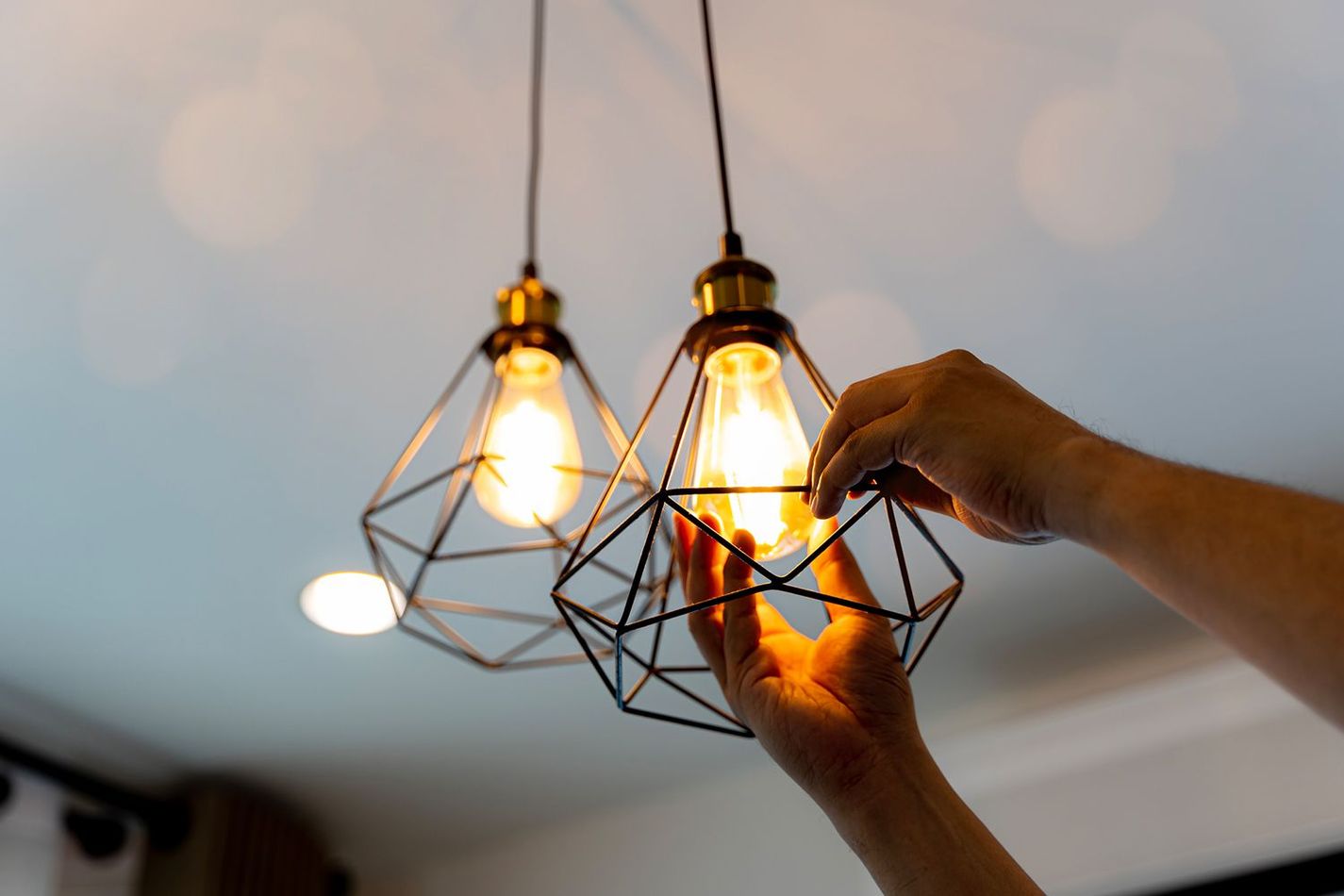 Person's hands adjusting a hanging pendant light with a geometric cage design, near another similar light on a white ceiling.