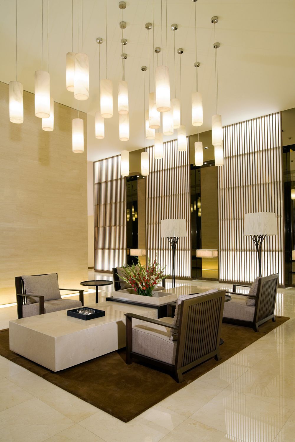 Elegant hotel lobby with seating area, pendant lights, marble floors, and patterned wall panels. Elegant hotel lobby with seating area, pendant lights, marble floors, and patterned wall panels.