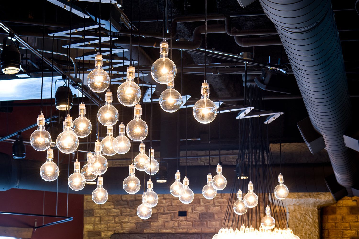 Cluster of glowing globe-shaped light bulbs hanging from a dark ceiling; brick and red wall background. Cluster of glowing globe-shaped light bulbs hanging from a dark ceiling; brick and red wall background.