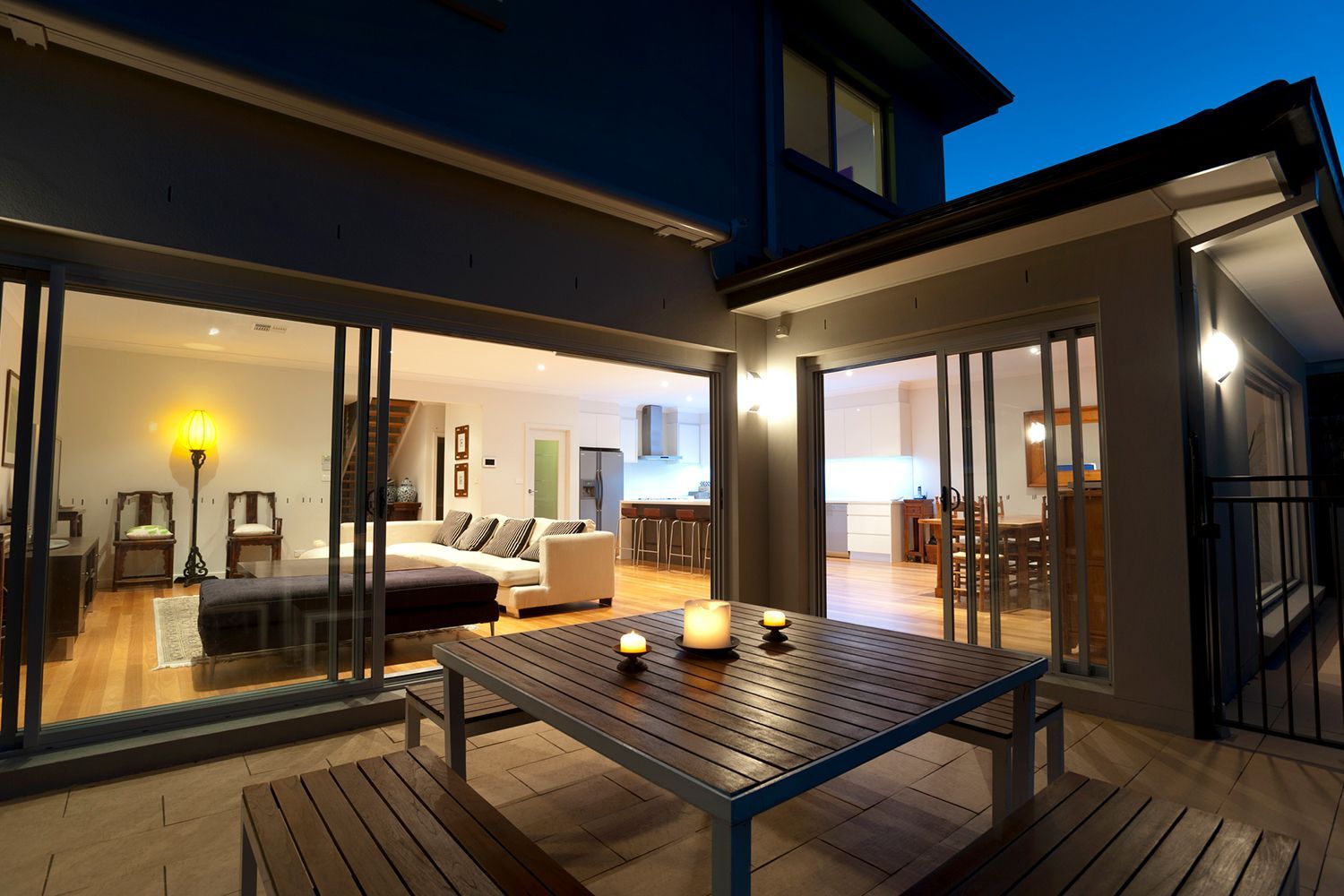 Patio with wooden table and benches, open to lit living and dining areas at dusk. Patio with wooden table and benches, open to lit living and dining areas at dusk.