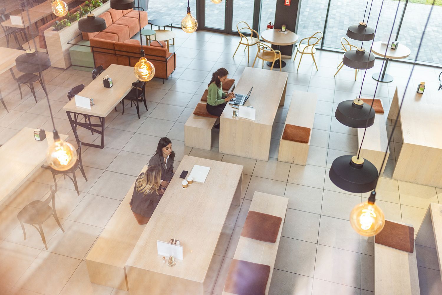 High-angle view of a modern cafe: people working at tables, warm lighting, wooden furniture, and large windows. High-angle view of a modern cafe: people working at tables, warm lighting, wooden furniture, and large windows.