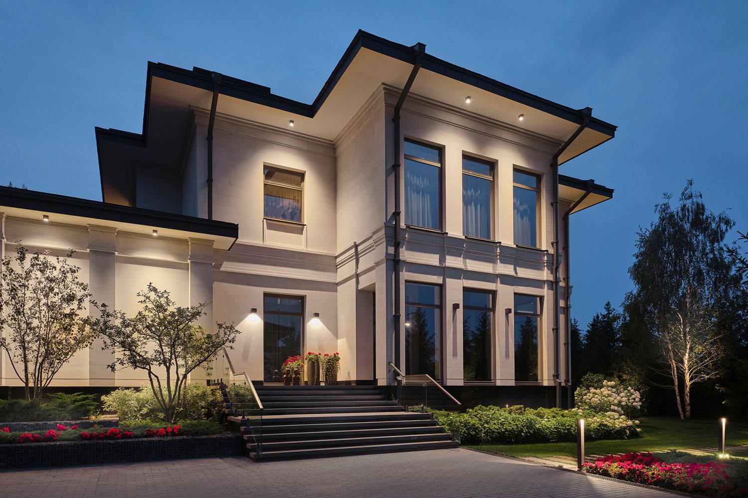 A modern two-story house illuminated at night. Features dark windows, outdoor lights, and manicured landscaping. A modern two-story house illuminated at night. Features dark windows, outdoor lights, and manicured landscaping.