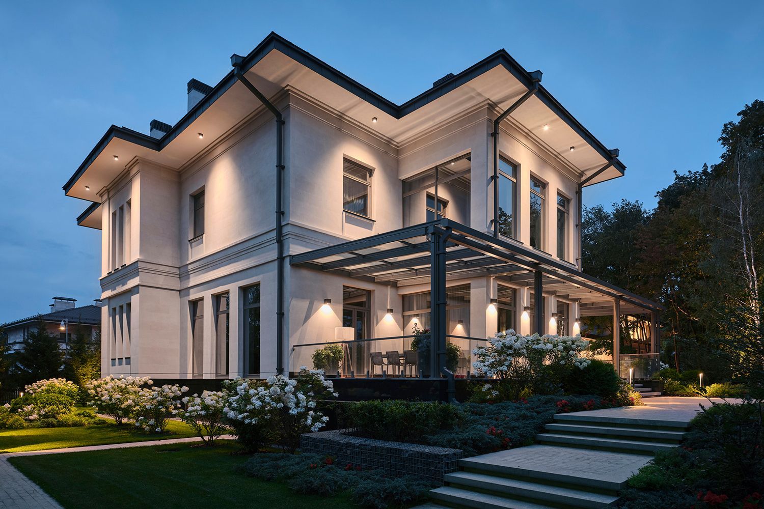 Two-story house with illuminated exterior, white walls, dark roof, porch, and landscaped garden at dusk. Two-story house with illuminated exterior, white walls, dark roof, porch, and landscaped garden at dusk.