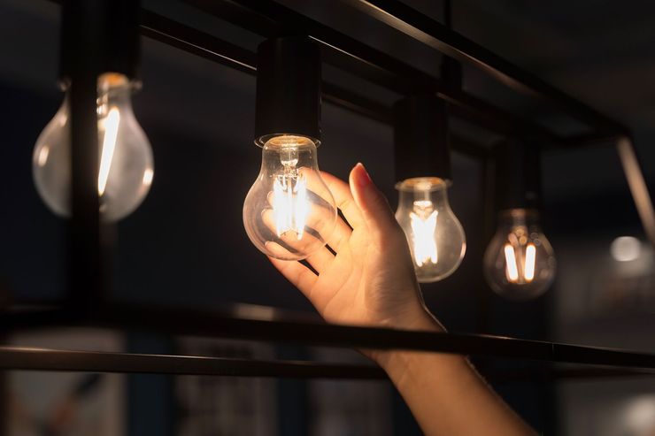 Hand reaching to touch a lit lightbulb in a row of light fixtures. Dark setting.