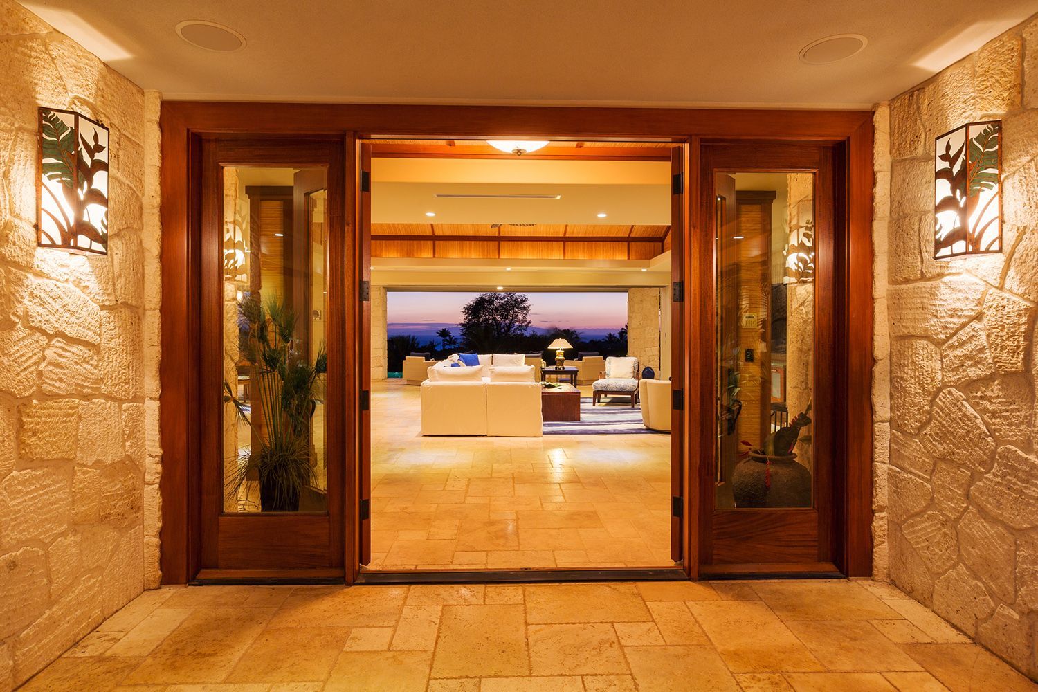Entrance with wooden doors and sidelights, stone walls, and warm lighting leading to a brightly lit living room.