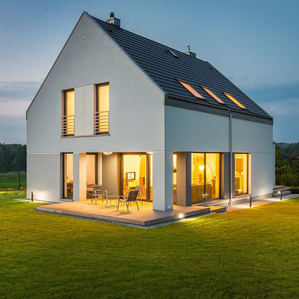 Modern two-story white house with a dark roof and illuminated windows, with lawn and patio in dusk. Modern two-story white house with a dark roof and illuminated windows, with lawn and patio in dusk.