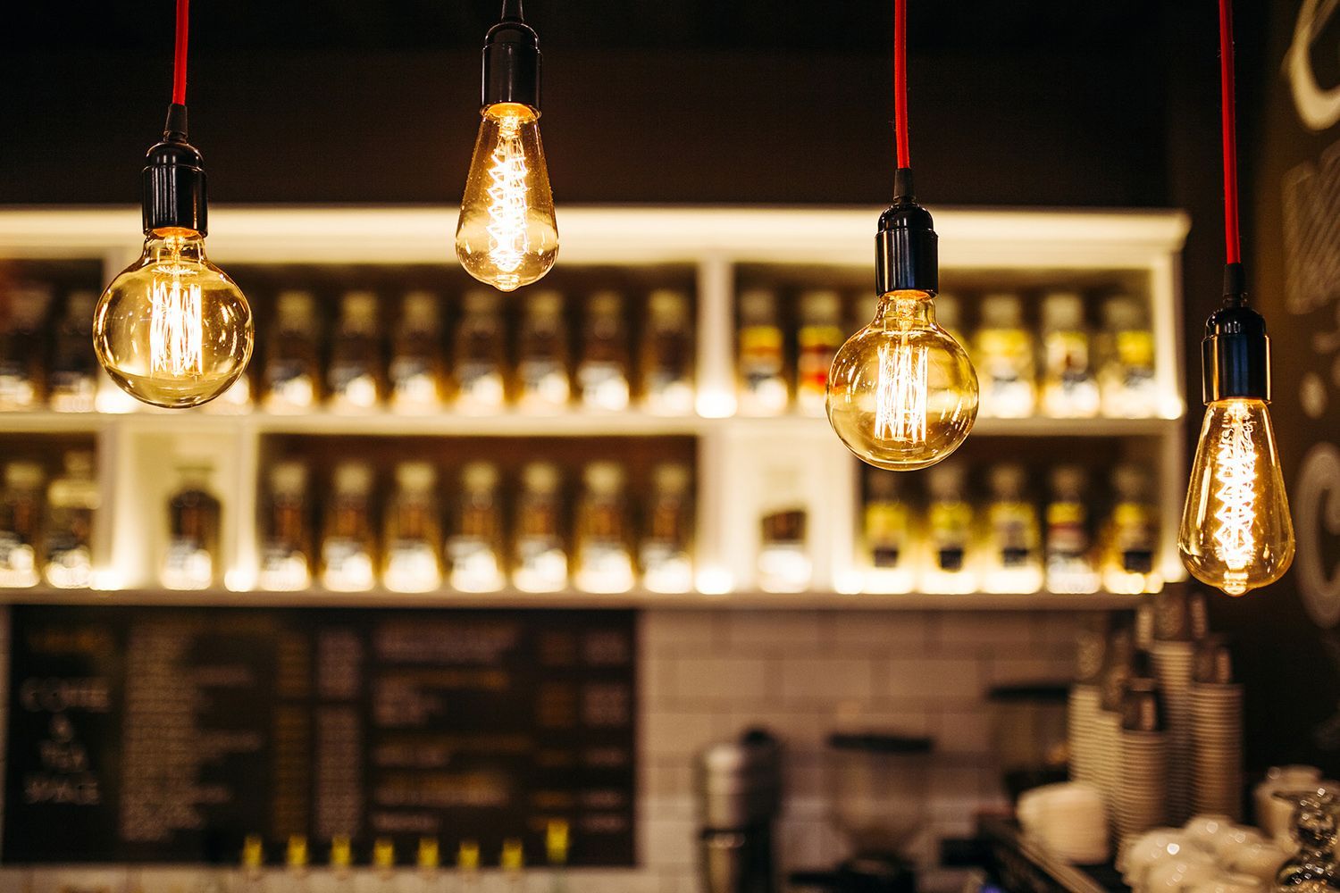 Four Edison-style light bulbs hang in a cafe, with shelves of jars and a menu board in the background. Four Edison-style light bulbs hang in a cafe, with shelves of jars and a menu board in the background.