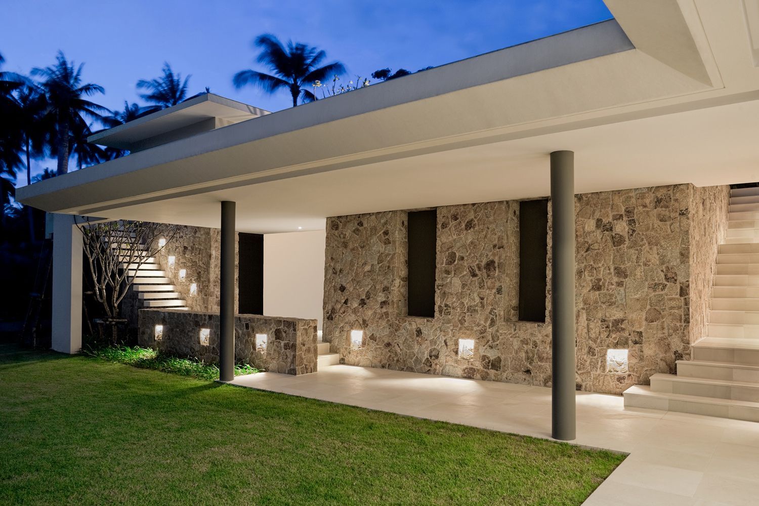 Modern house exterior with stone wall, pillars, and lit stairs, at dusk.