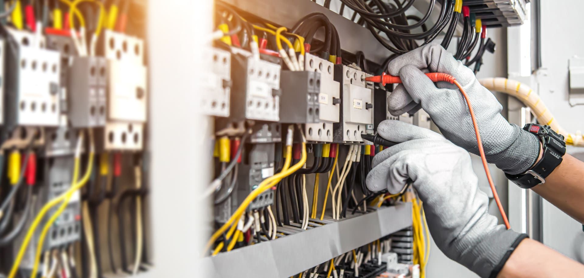 An electrician in grey gloves tests wires in an electrical panel with a multimeter. Yellow and black wires are visible.