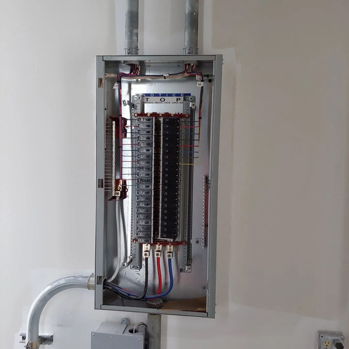 Open electrical panel with wires and circuit breakers, mounted on a white wall.