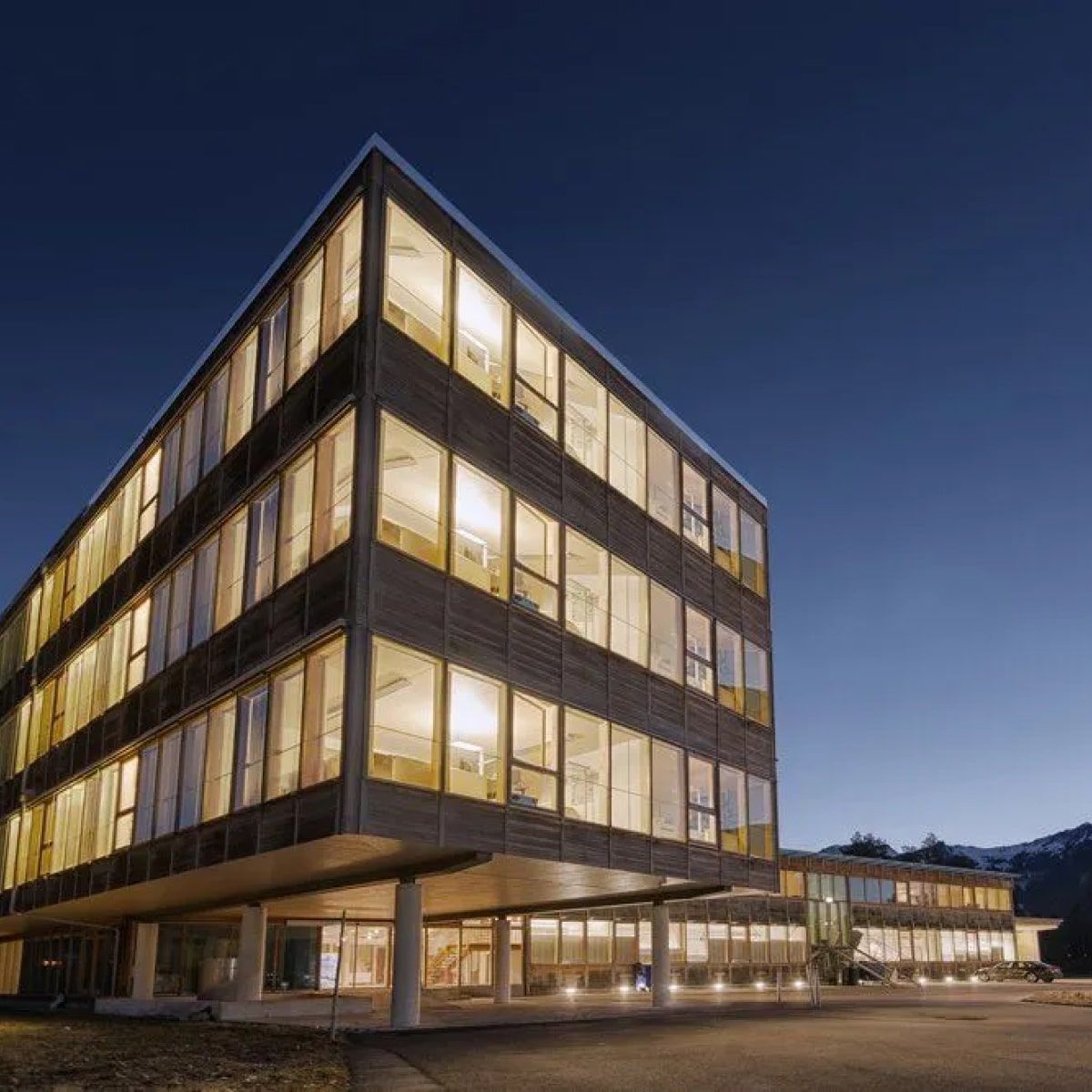 Modern building with multiple lit floors at dusk. White pillars support the upper floors, with a clear sky in the background.